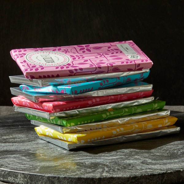 5 colorfully wrapped Xocolatl chocolate bars in the colors of bright yellow, grass green, cherry red, teal blue & light pink are stacked on top of each other interspersed with bars wrapped only in silver foil.