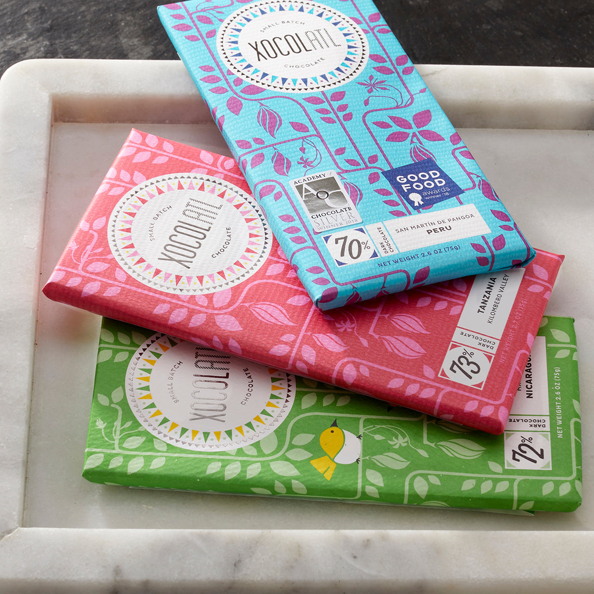 3 Xocolatl chocolate bars wrapped in green, cherry red & teal blue paper wrappers fanned out on a marble platter.