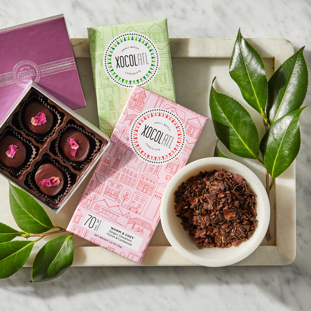 An open 4 piece box of chocolate bonbons topped with rose petals on a white marble tray with 2 Xocolatl Chocolate bars wrapped in pink and green and a bowl of cacao husk tea.