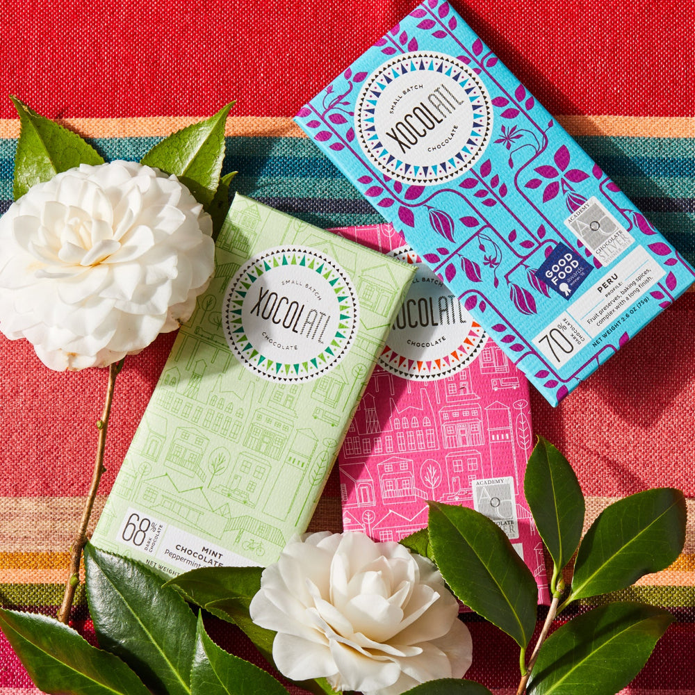 3  Xocolatl chocolate bars wrapped in light green, hot pink, and teal blue wrappers and two white flowers all lying on colorful fabric.