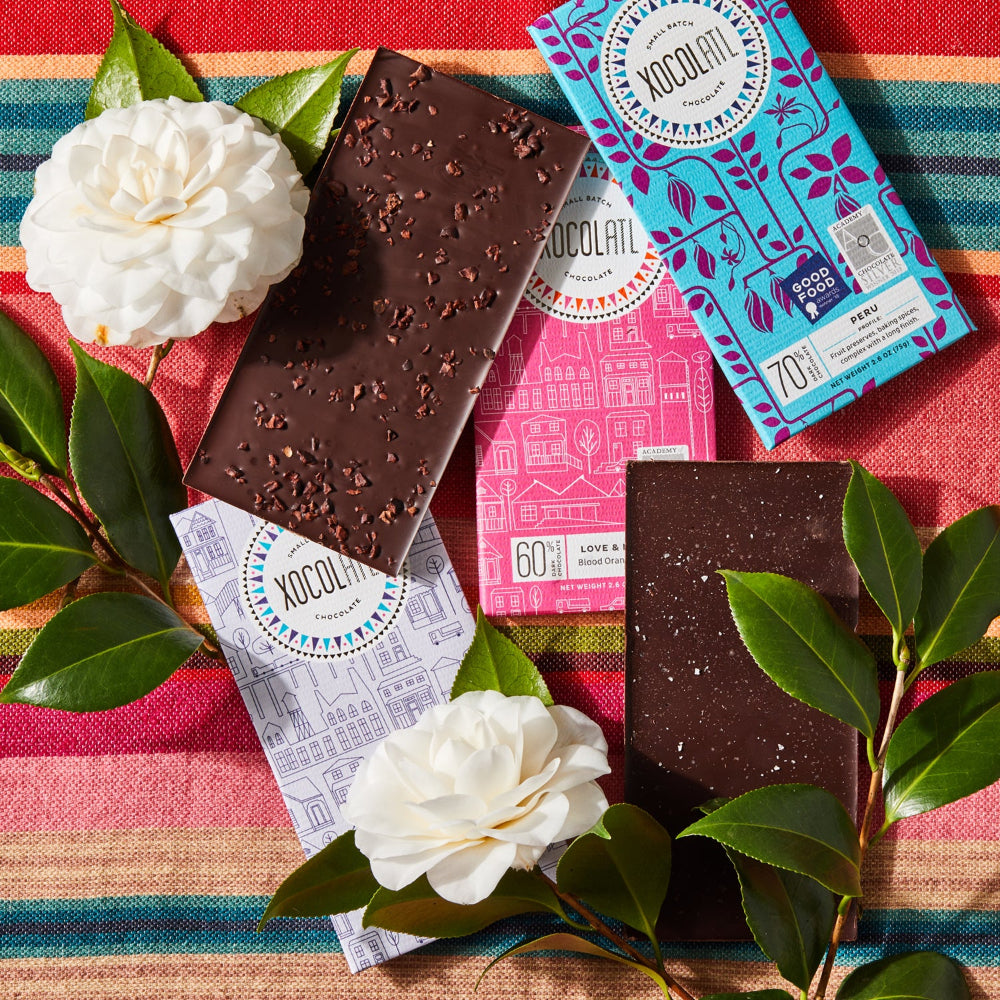 3  Xocolatl chocolate bars wrapped in light purple, hot pink, and teal blue wrappers lying with 2 unwrapped chocolate bars and two white flowers on colorful fabric.
