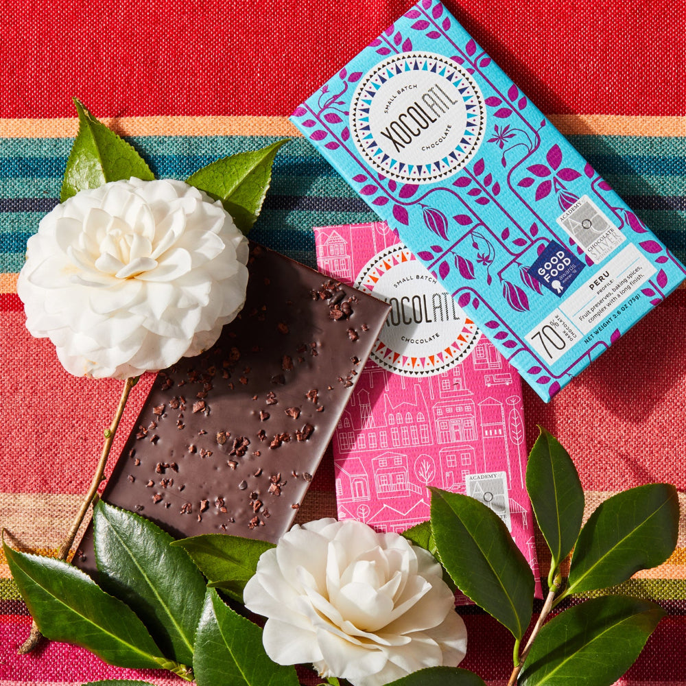 2  Xocolatl chocolate bars wrapped in hot pink and teal blue wrappers and one unwrapped chocolate bar showing a sprinkle of cocoa nibs, lying with white flowers on colorful fabric.