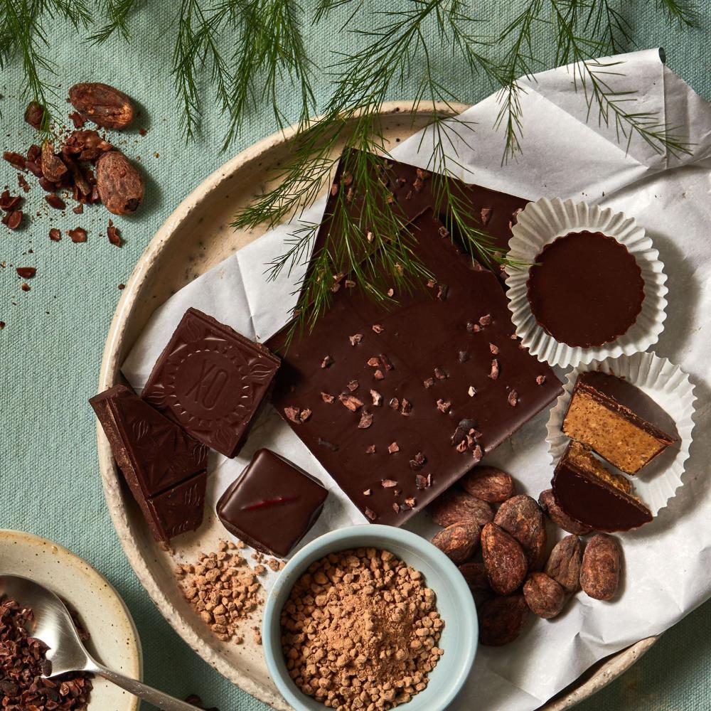 The contents of a small chocolate subscription are plated on a round, cream plate, showing 2 unwrapped Xocolatl Chocolate bars, 3 chocolate bonbons ready to be eaten and several whole cacao beans.