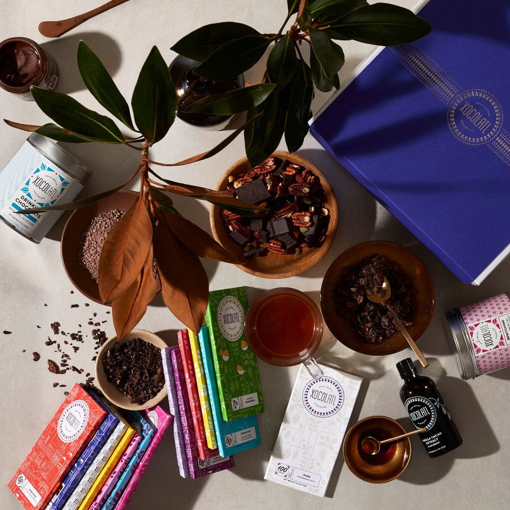 A variety of wrapped & unwrapped Xocolatl Chocolate products are displayed on a white table top along with a royal blue Xocolatl branded gift box & decorative magnolia branches. There are 2 stacks of chocolate bars in rainbow hued wrappers, bowls of chocolate trail mix & baking chocolate, along with tins of Xocolatl tea & containers of chocolate hazelnut spread, cacao extract & drinking chocolate.