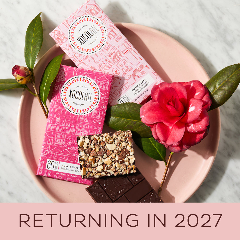 A set of three chocolate bars presented on a plate with a pink flower and green leaves in the background. Each bar has a unique topping, and the set is tied together with a silver paper belly-band.