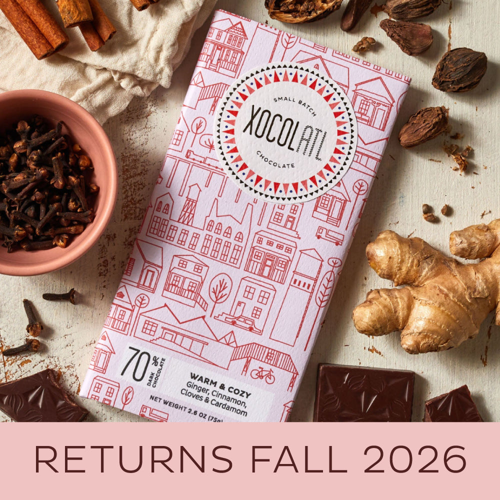 A Xocolatl Chocolate bar in light pink packaging called Warm and Cozy on a light surface lying beside fresh ginger, cinnamon sticks, Cardamom pods and cloves.
