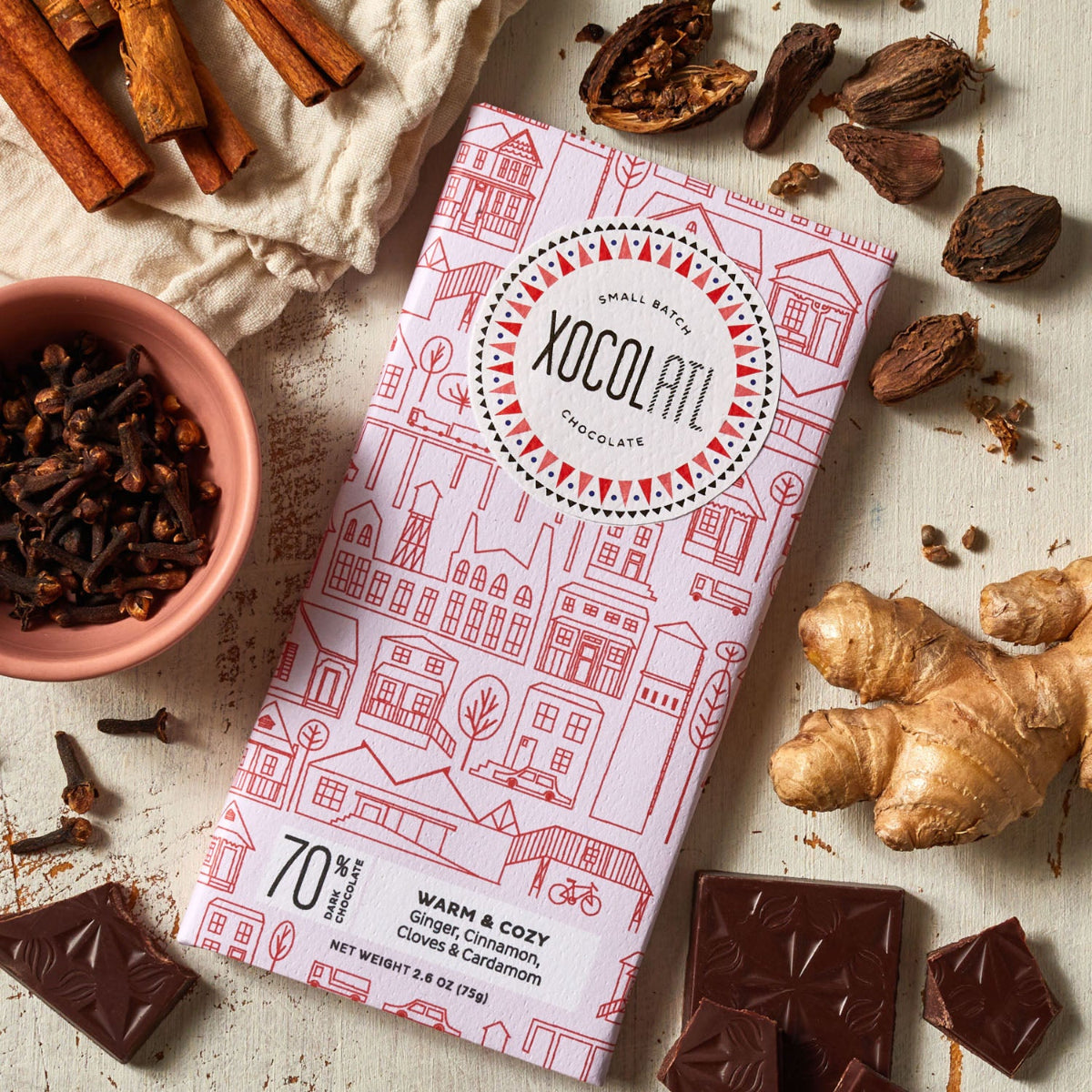 A Xocolatl Chocolate bar in light pink packaging called Warm and Cozy on a light surface lying beside fresh ginger, cinnamon sticks, Cardamom pods and cloves.