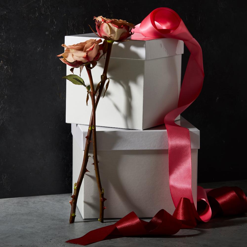 One small, two-piece, lidded white gift box is stacked on top of a larger white gift box with a spool of pink satin ribbon unfurled around them and a rose for decoration.