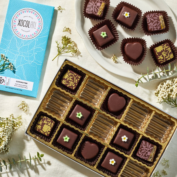 An open 15 piece box of variously decorated chocolate bonbons with a teal blue wrapped Xocolatl Chocolate bar of the flavor, "Commuter" on a white table with small white flowers.