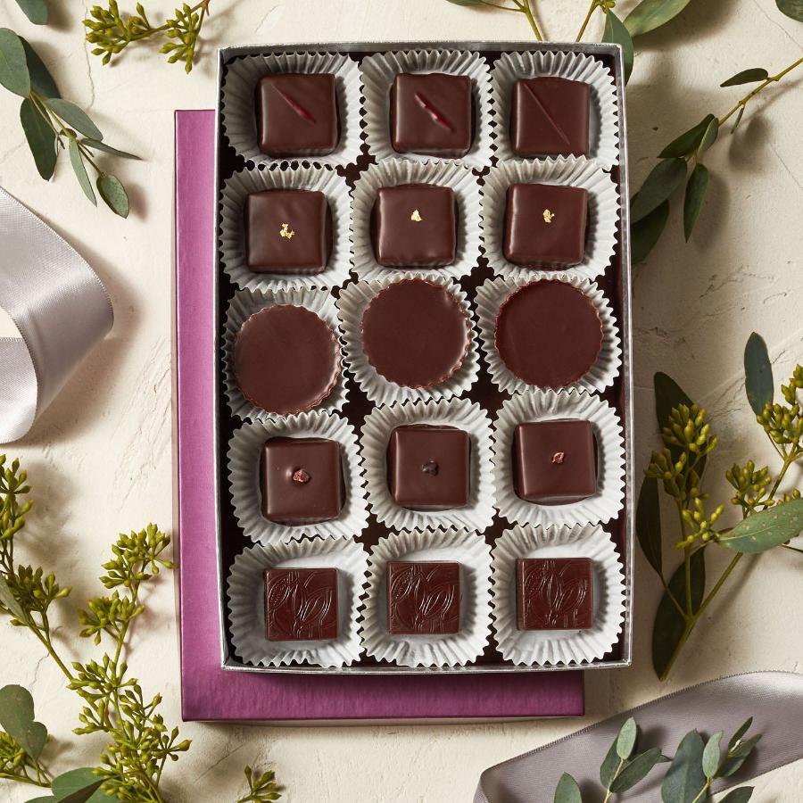 An open box of 15 Xocolatl Chocolate bonbons are set on a white table top with decorative foliage. Inside the box are 5 rows of 3 bonbons each. The 3 bonbons in each row are identical while the 5 bonbons in each column are different, indicating 3 bonbons each of 5 different flavors.