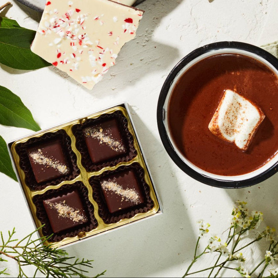 A white table set with holiday greenery, a plate holding pieces of white & dark chocolate peppermint bark, a mug of hot chocolate with a marshmallow, and an opened box with 4 chocolate bonbons.