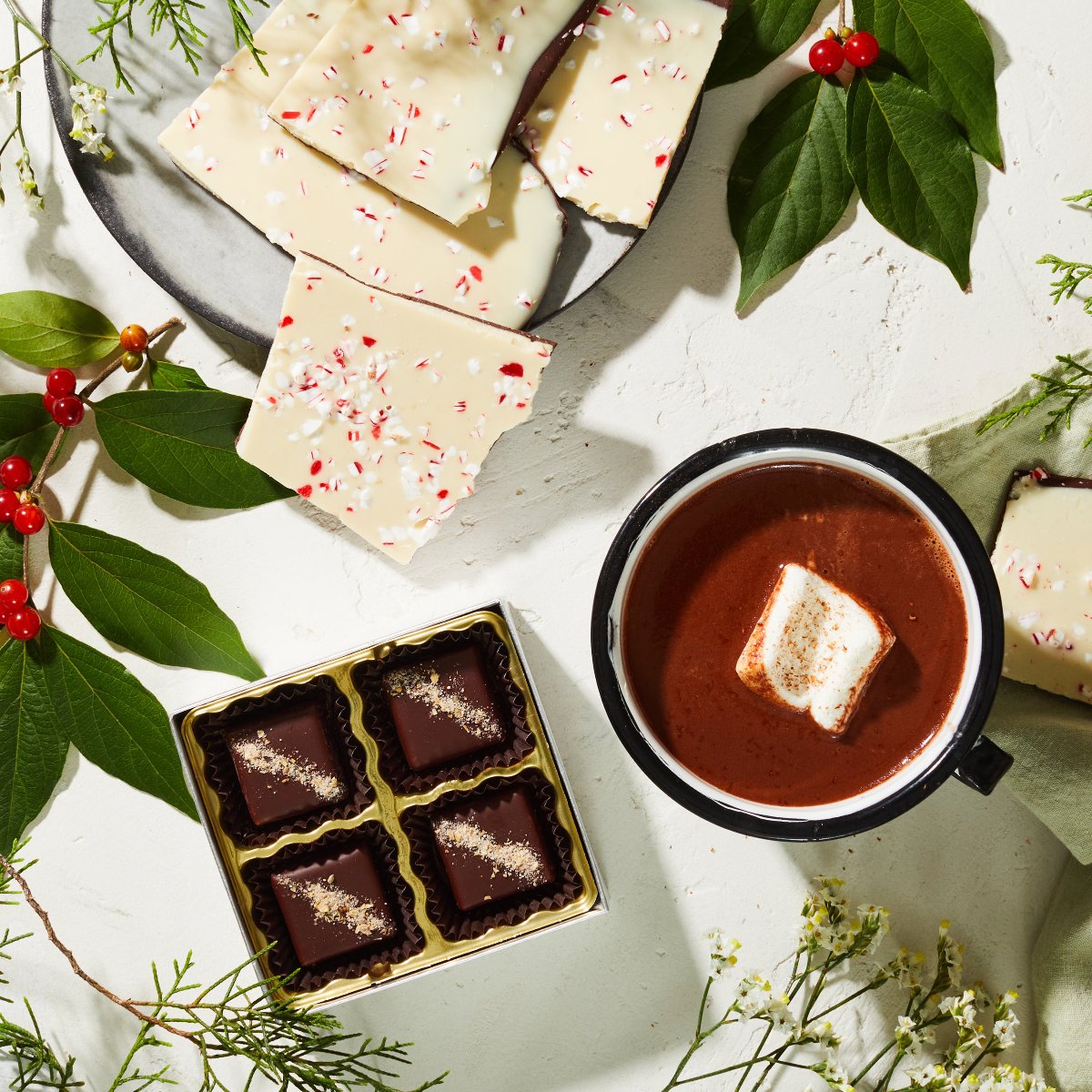 A white table set with holiday greenery, a plate holding pieces of white & dark chocolate peppermint bark, a mug of hot chocolate with a marshmallow, and an opened box with 4 pieces of square chocolate caramels.