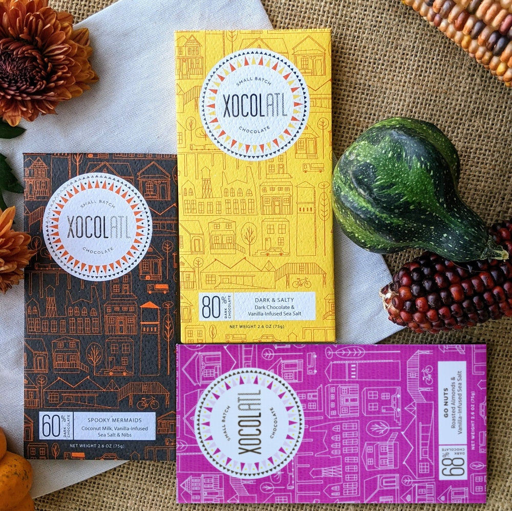 Three Xocolatl chocolate bars (Spooky Mermaids, Dark & Salty and Go Nuts!) on burlap and a white napkin with orange mums, a gourd and ornamental corn.