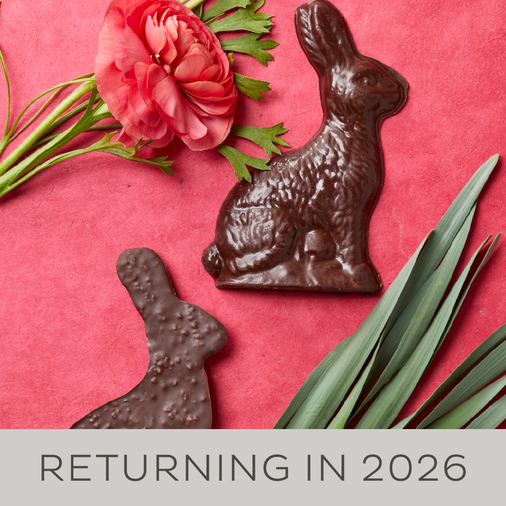 One chocolate rabbit shown in profile is face up and one is face down displaying the bumpy puffed crisps mixed into the chocolate. They are displayed on a red background with a red ranunculus bloom. A banner along the bottom reads "Returning in 2026".