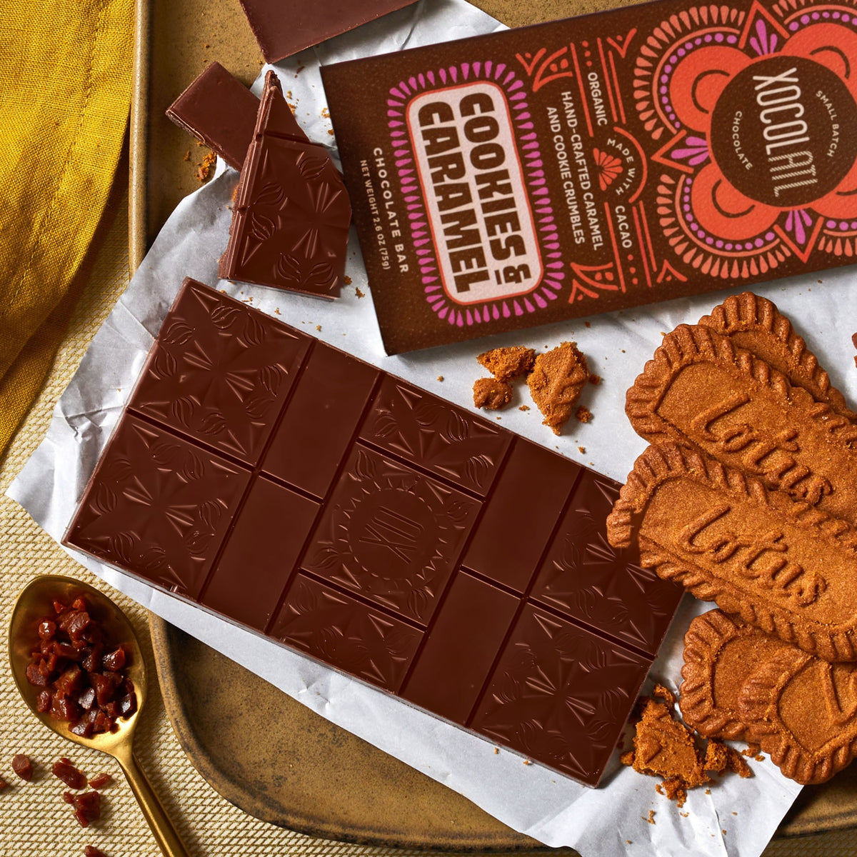 A brown Cookies & Caramel wrapped Xocolatl chocolate bar beside an unwrapped bar and broken pieces, with Biscoff cookies, cookie crumbles and a gold spoon of caramel pieces.