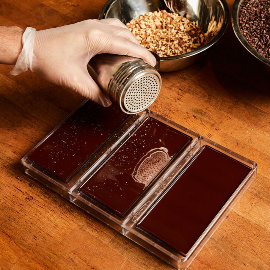 A Xocolatl Chocolate maker's gloved hand shakes white chunks of sea salt out of a silver shaker and on to the backs of 3 Xocolatl Chocolate bars. A bowl of nuts and a bowl of cacao nibs are in the background.