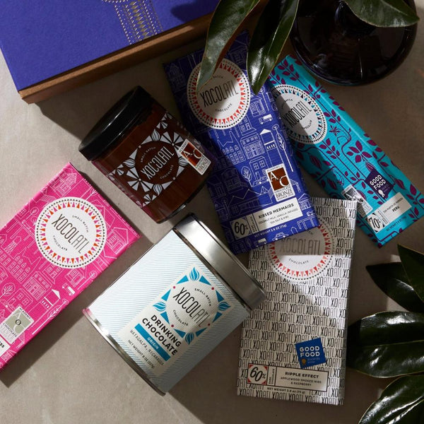 On a white table, 4 Xocolatl chocolate bars are arrayed, in hot pink, royal blue, teal, & white wrappers. Interspersed with the bars is a glass jar of chocolate hazelnut spread, a blue & white tin of drinking chocolate, & the corner of a royal blue Xocolatl gift box.