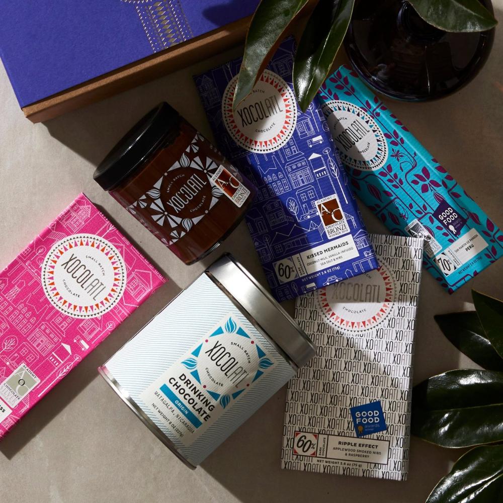 On a white table, 4 Xocolatl chocolate bars are arrayed, in hot pink, royal blue, teal, & white wrappers. Interspersed with the bars is a glass jar of chocolate hazelnut spread, a blue & white tin of drinking chocolate, & the corner of a royal blue Xocolatl gift box.
