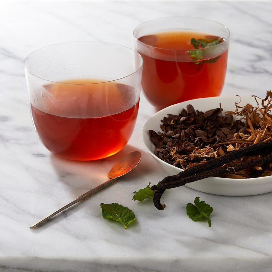 Two clear glasses of herbal cacao tea on a marble surface, with a bowl of cacao husks and a spoon with mint leaves and vanilla pods nearby.
