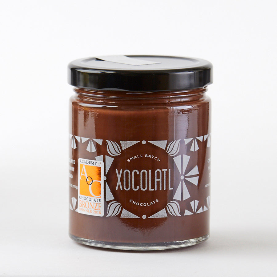 A glass jar with a black lid on a white background. The jar says Xocolatl Chocolate and is filled with a chocolate hazelnut paste inside. There is a bronze, Academy of Chocolate award sticker on the jar.