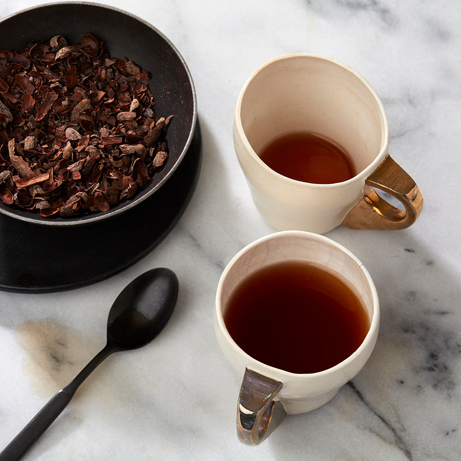 Two cups of herbal cacao tea on a marble surface with a bowl of roasted cacao husks and nibs in the background.
