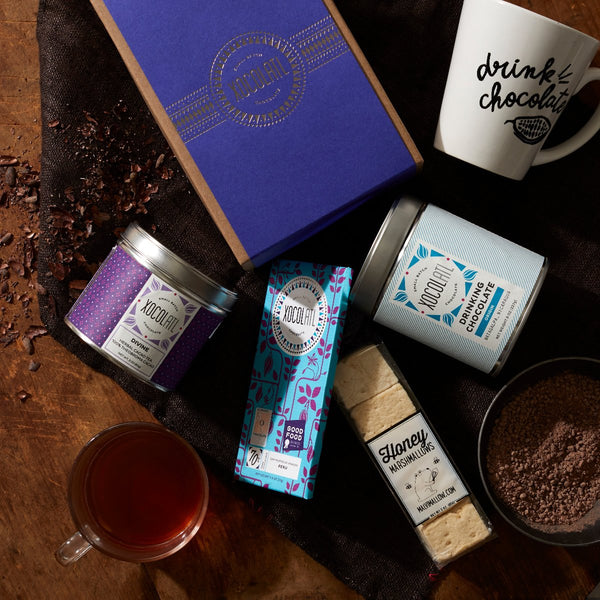 A purple tin of Xocolatl "Divine" herbal cacao tea, lying beside a teal-wrapped Xocolatl "Peru 70% Dark" chocolate bar, a blue & white tin of Xocolatl "Drinking Chocolate", a pouch of Honey flavored marshmallows & a white mug with "drink chocolate" printed in black on a wooden tabletop.