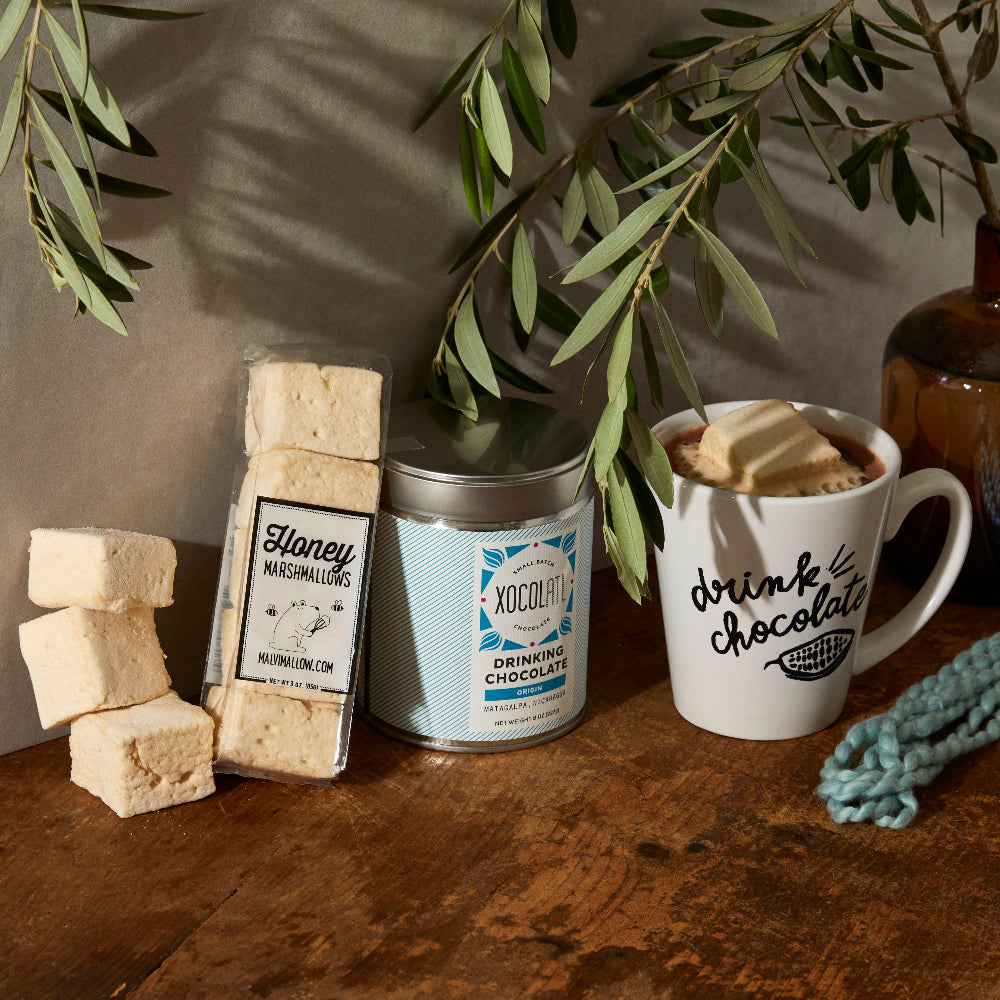 The image features a delightful Drinking Chocolate Bundle, ideal for enhancing your hot cocoa experience. The bundle includes a mug with a playful cacao pod design, a stand-up pouch of rich, European-style drinking chocolate made from decadent, single-origin Nicaraguan dark chocolate and organic cane sugar, and a pack of fluffy, honey-flavored Honey Marshmallows.
