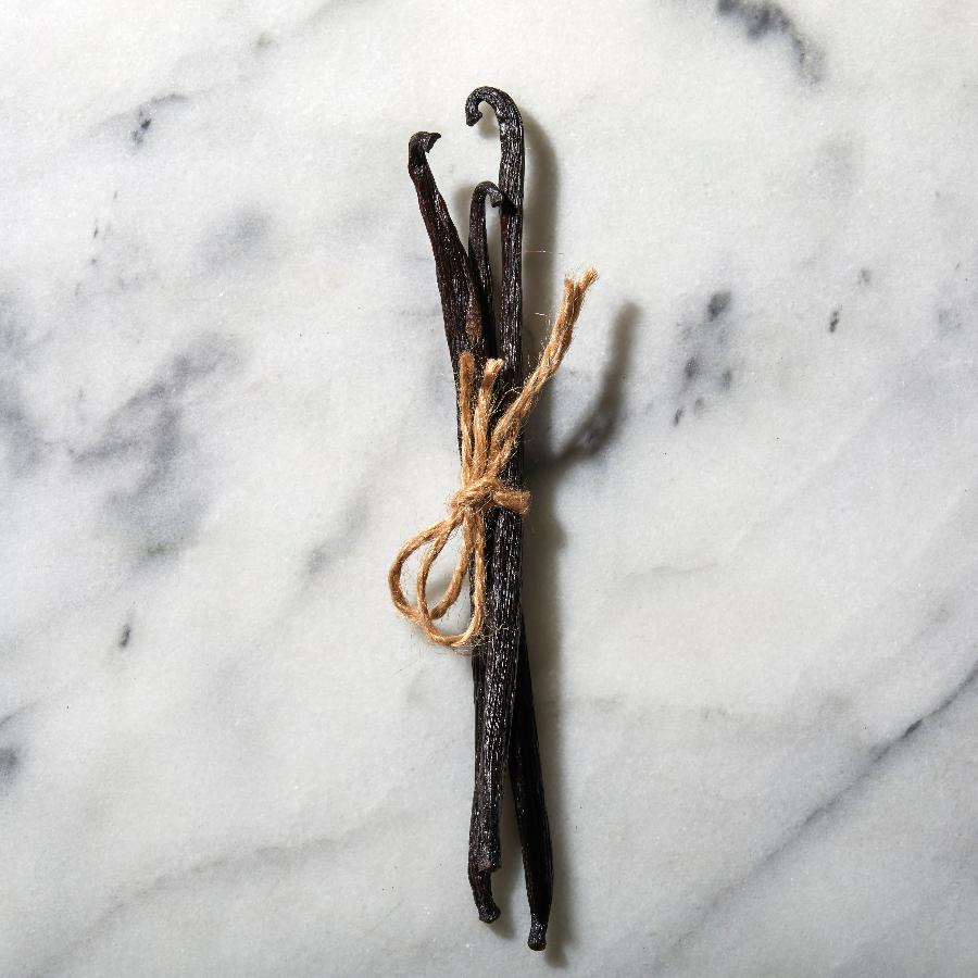 3 long, slender, black-brown vanilla pods are bundled together, tied with a piece of brown twine upon a white marble surface. Each pod curls at the top tip like a swan peering down.