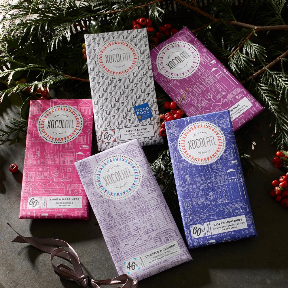 5 Xocolatl chocolate bars in pink, lavender, royal blue , plum, & black & white paper wrappers laying under festive greenery. They are the flavors "Love & Happiness", "Crackle & Crunch" and "Kissed Mermaids", "Go Nuts", & "Ripple Effect".