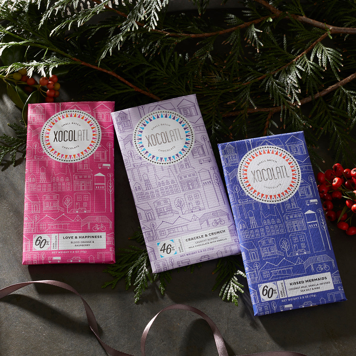 3 Xocolatl chocolate bars in pink, lavender & royal blue paper wrappers laying under festive greenery. They are the flavors "Love & Happiness", "Crackle & Crunch" and "Kissed Mermaids".