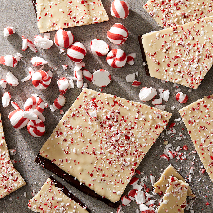 A close up of 9 pieces of peppermint bark clustered on a grey table top with crushed, red & white peppermint candies. The bark is a layer of dark chocolate, covered in a layer of cream colored white chocolate, with a layer of red & white candy pieces.