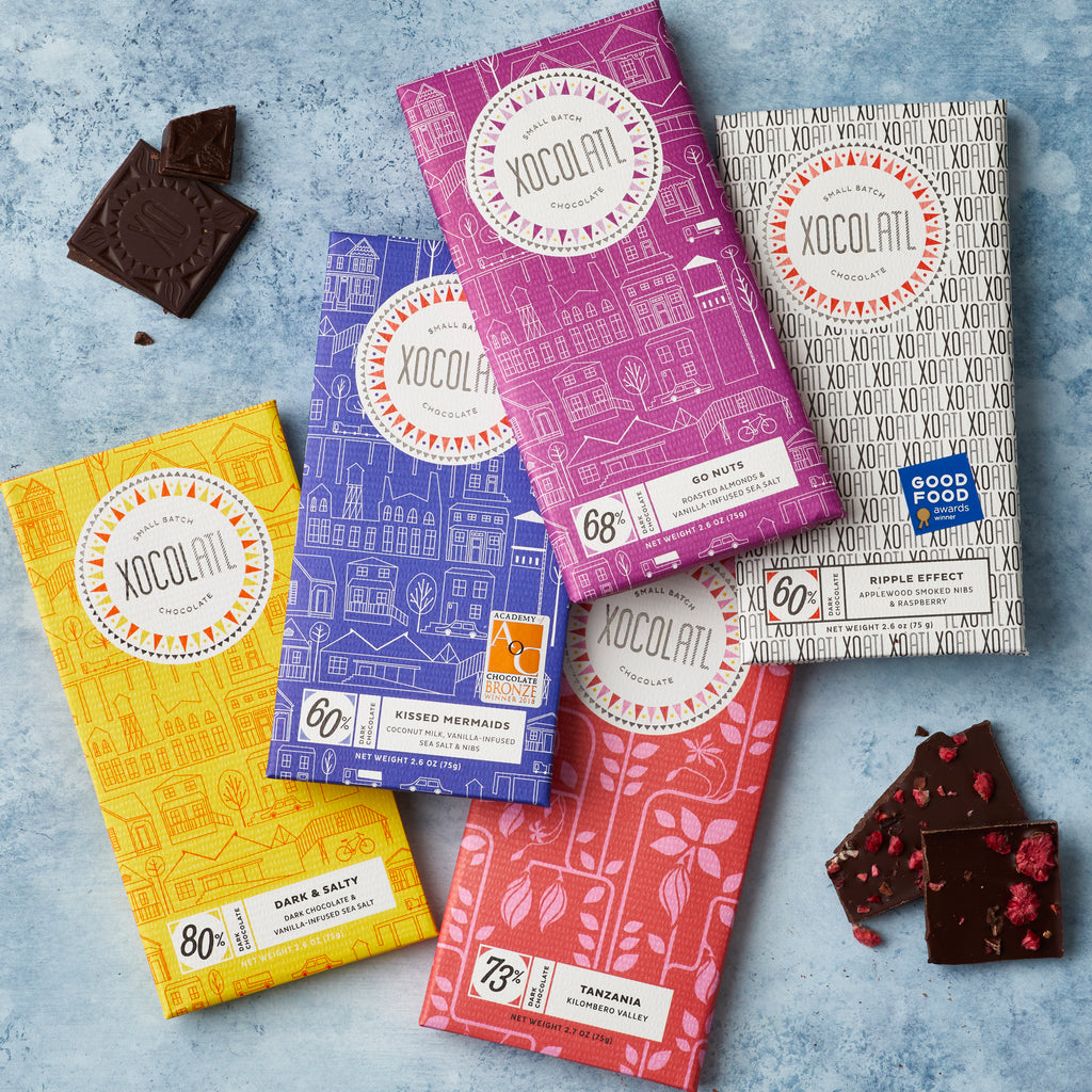 5 Xocolatl chocolate bars on a light blue background in yellow, blue, purple, red, and white wrappers beside a squares of chocolate with the XO logo and raspberries.