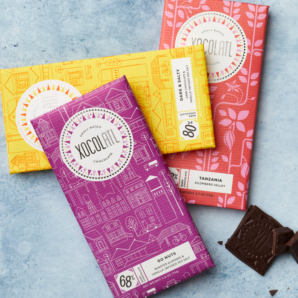 3 Xocolatl chocolate bars on a light blue background in purple, yellow and red wrappers beside a square of chocolate with the XO logo.