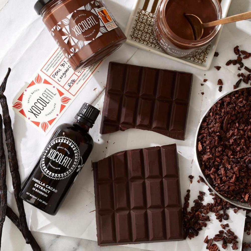 3 loose vanilla beans, an amber bottle of Xocolatl Vanilla Cacao Extract, 2 glass jars of Xocolatl Hazelnut spread (one closed, one open with a spoon), a bowl of nibs & a thick slab of baking chocolate lay together on a white countertop, ready for immediate use in a recipe.