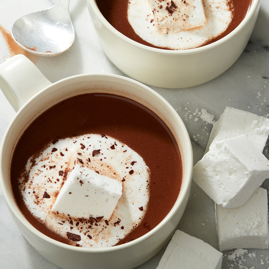 2 white mugs on a white marble slab, each filled almost to the brim with dark brown hot chocolate. In the center of each mug is a melting marshmallow, its edges spreading out  from the marshmallow body like an airy, twirling white skirt.