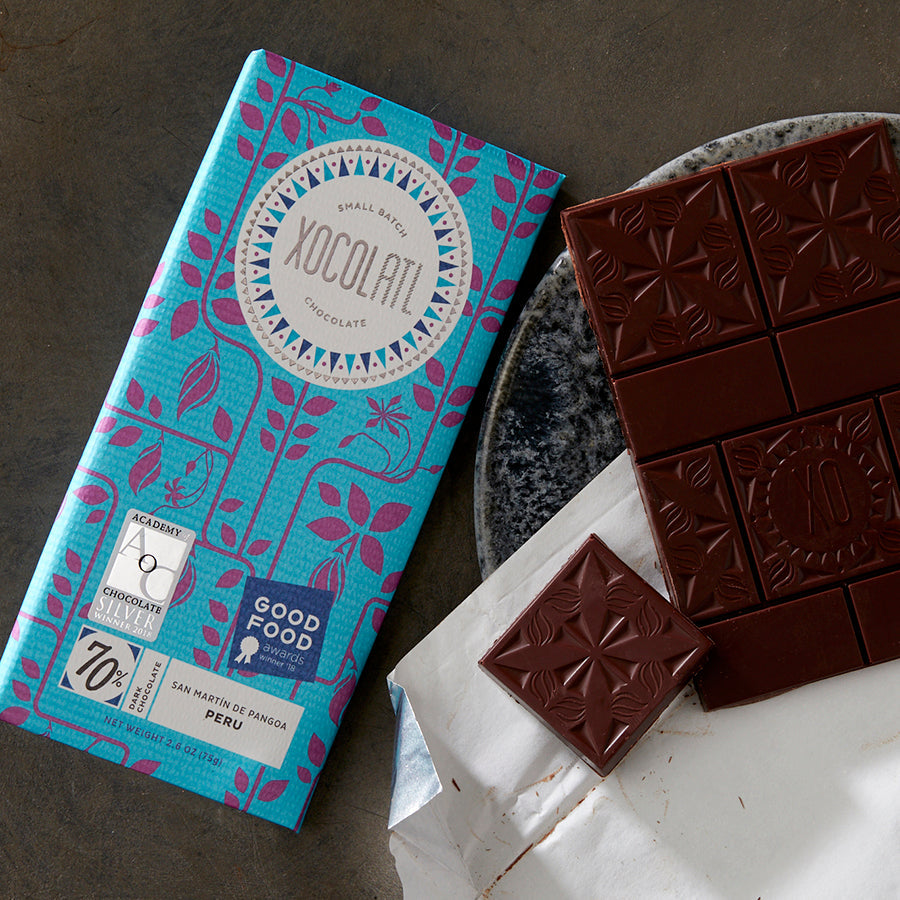 A teal blue Xocolatl chocolate bar of the flavor "70% Single Origin Peru" beside an unwrapped and partially eaten chocolate bar on a grey, stone plate.